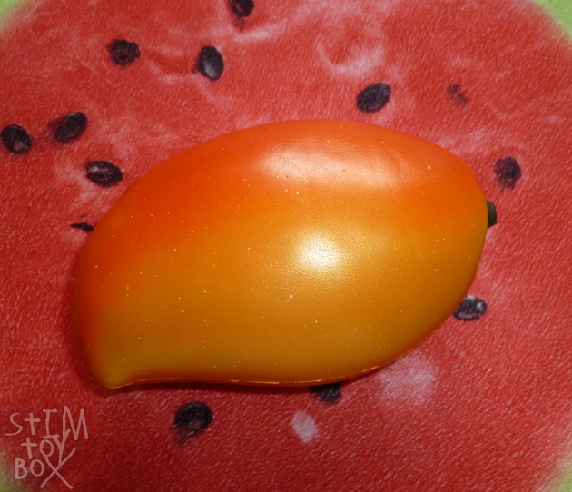 A photo of an orange mango squishy sitting on a red watermelon slice pillow. The squishy is shaped like a mango with a brown stem and a two-tone light-orange to dark orange gradient effect, splattered with fine white paint-speckle dots. It’s covered with a shiny slightly-opalescent paint that reflects the camera’s flash.
