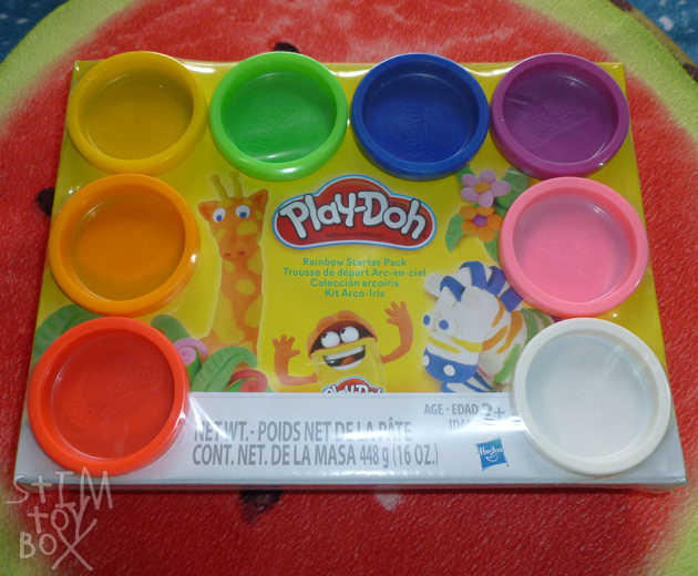 Play-Doh’s Rainbow Starter Pack, a selection of eight yellow plastic tubs, topped with a matching coloured plastic lid, of red, orange, yellow, green, blue, purple and pink dough. Photo shows the tubs inside a box and covered with cling film, surrounding a picture of a Play-Doh zebra and giraffe surrounded by leaves and flowers in the centre of the box. 